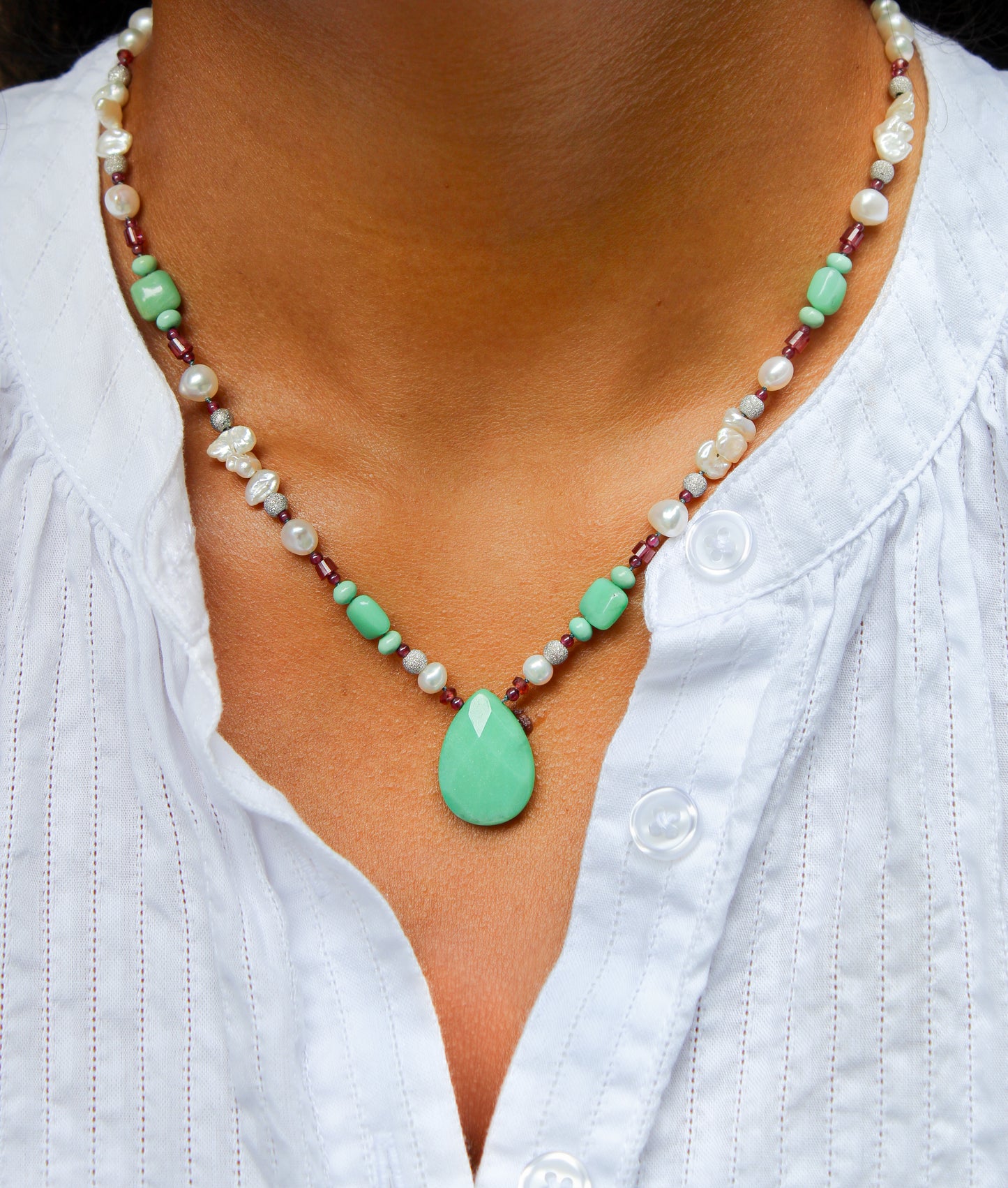 A close-up of a person wearing a beaded necklace with a mint green teardrop pendant, white pearl-like beads, and small green beads over a white button-up shirt.