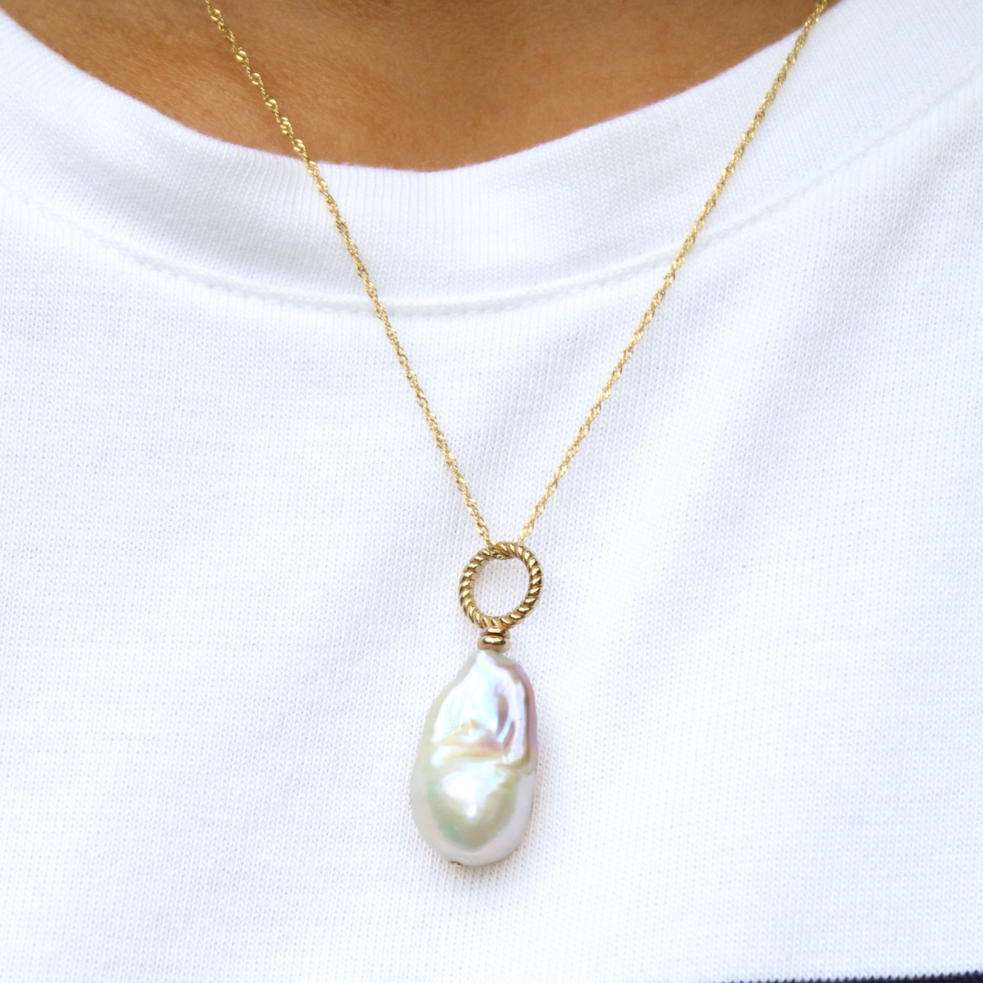 A gold chain necklace with a baroque pearl pendant, worn by a person wearing a white t-shirt with black horizontal stripes.