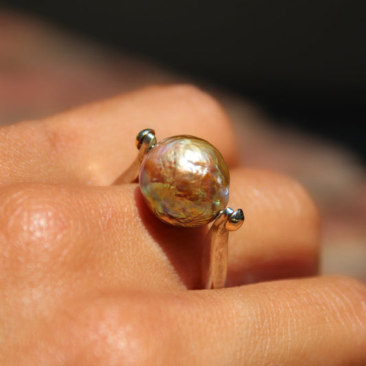 A close-up of a hand wearing a unique ring featuring a large, textured, iridescent gold-toned glass bead suspended on a thin silver-toned band.