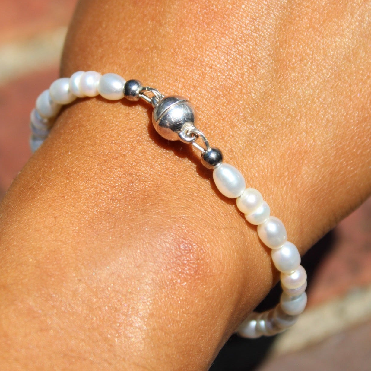 A close-up of a person's wrist wearing a beaded bracelet made of white pearl-like beads and a shiny silver ball accent, set against a blurred brick background.