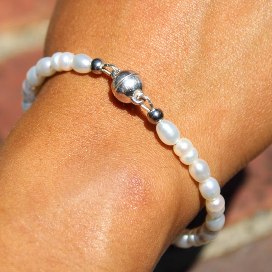 A close-up of a person's wrist wearing a beaded bracelet made of white pearl-like beads and a shiny silver ball accent, set against a blurred brick background.